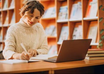 Uma mulher jovem, concentrada, usando um laptop e escrevendo em um caderno em uma biblioteca.