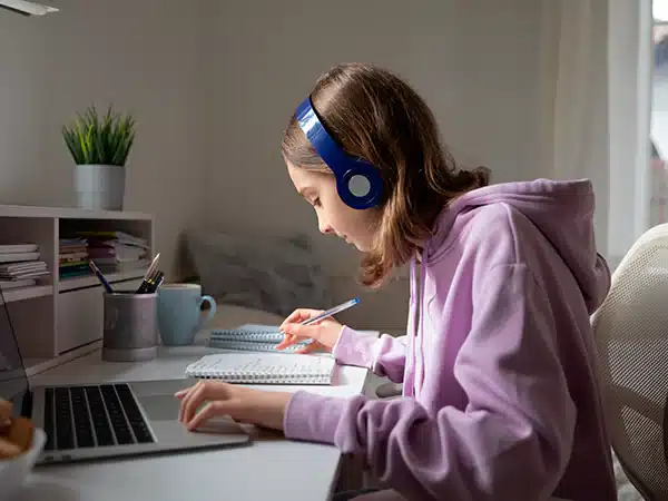 Pessoa estudando com fones de ouvido, concentrada em suas anotações, com um computador à sua frente, representando a relação entre estudo e música.
