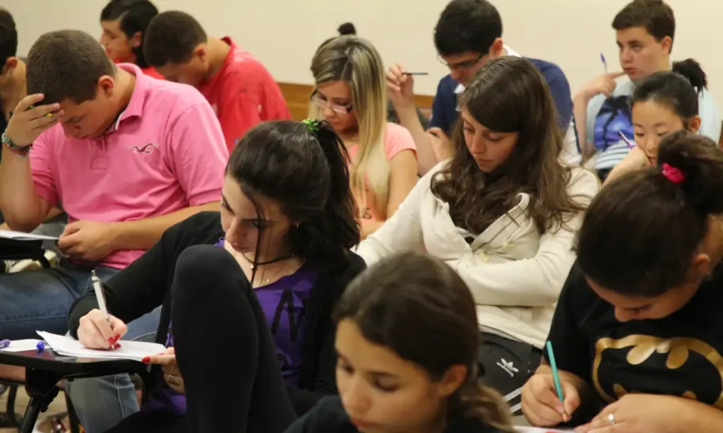 Grupo de estudantes em uma sala de aula, concentrados em seus cadernos enquanto se preparam para uma prova ou vestibular.