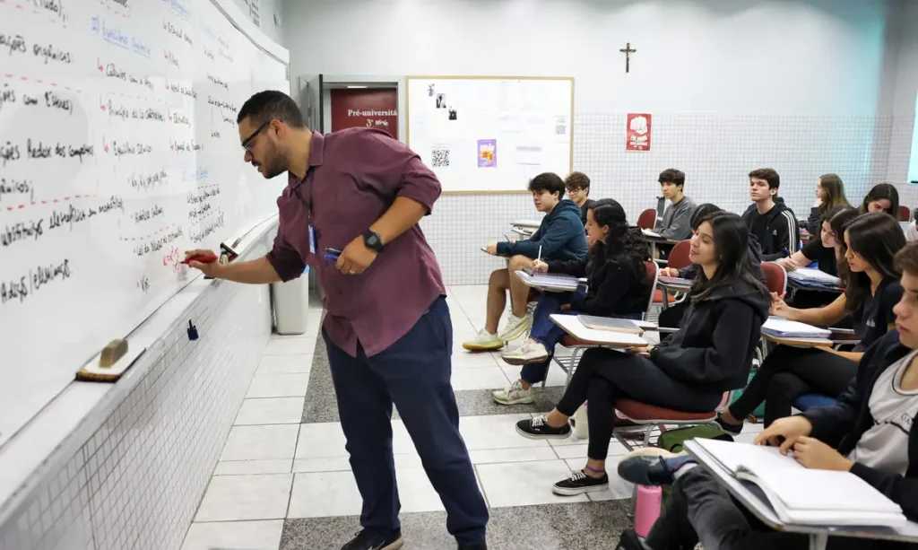 Correção de Desempenho: Preparação Docente e o Gabarito PND 2025 Professor de camisa vinho e óculos escrevendo em um quadro branco durante uma aula, com alunos sentados em fileiras assistindo e tomando notas em uma sala de aula preparatória.