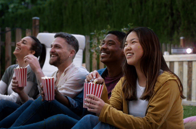 Grupo de amigos sentados no jardim com baldes de pipoca, sorrindo e assistindo filme ao ar livre