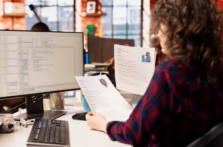 Recrutadora analisando currículos impressos e dados de candidatos no computador.