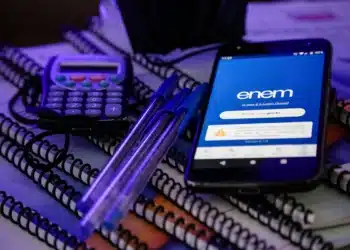 Um smartphone exibindo a tela de login do Enem, acompanhado de uma calculadora e canetas azuis sobre cadernos, simbolizando a preparação para o exame.