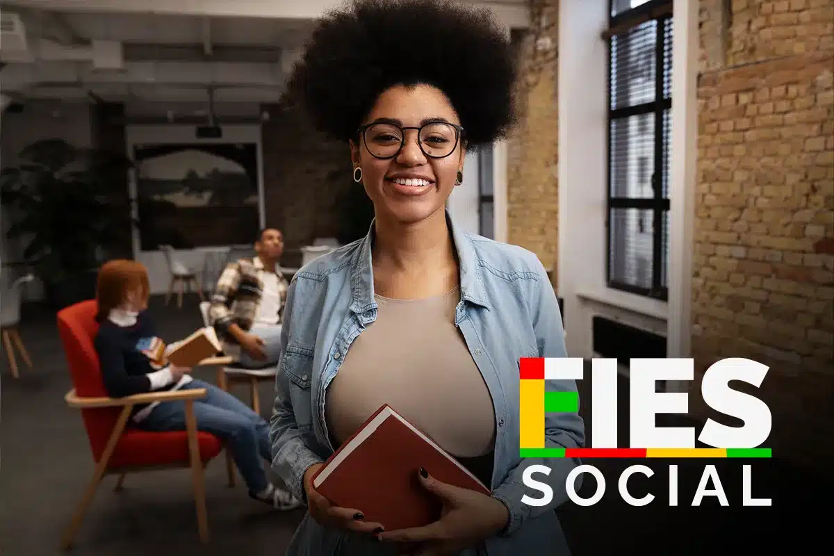 Jovem estudante feliz com livro no programa FIES Social Mulher sorridente segurando livro em ambiente de estudo com pessoas ao fundo e logo FIES Social
