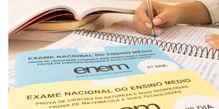 Mão escrevendo em um caderno de estudos com os cadernos de prova do Enem à frente, representando a preparação e os critérios de correção da redação.