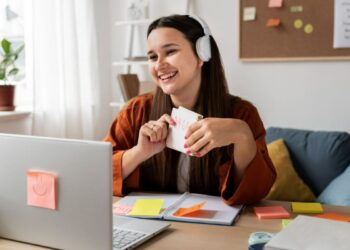 Jovem sorridente usando fones de ouvido em frente a um notebook, segurando cartões de estudo, representando o aprendizado de IA por meio de cursos.