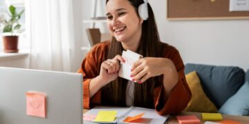 Jovem sorridente usando fones de ouvido em frente a um notebook, segurando cartões de estudo, representando o aprendizado de IA por meio de cursos.
