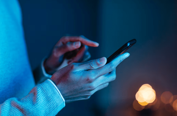 Mãos de pessoa usando smartphone com iluminação em tons azuis e fundo desfocado à noite