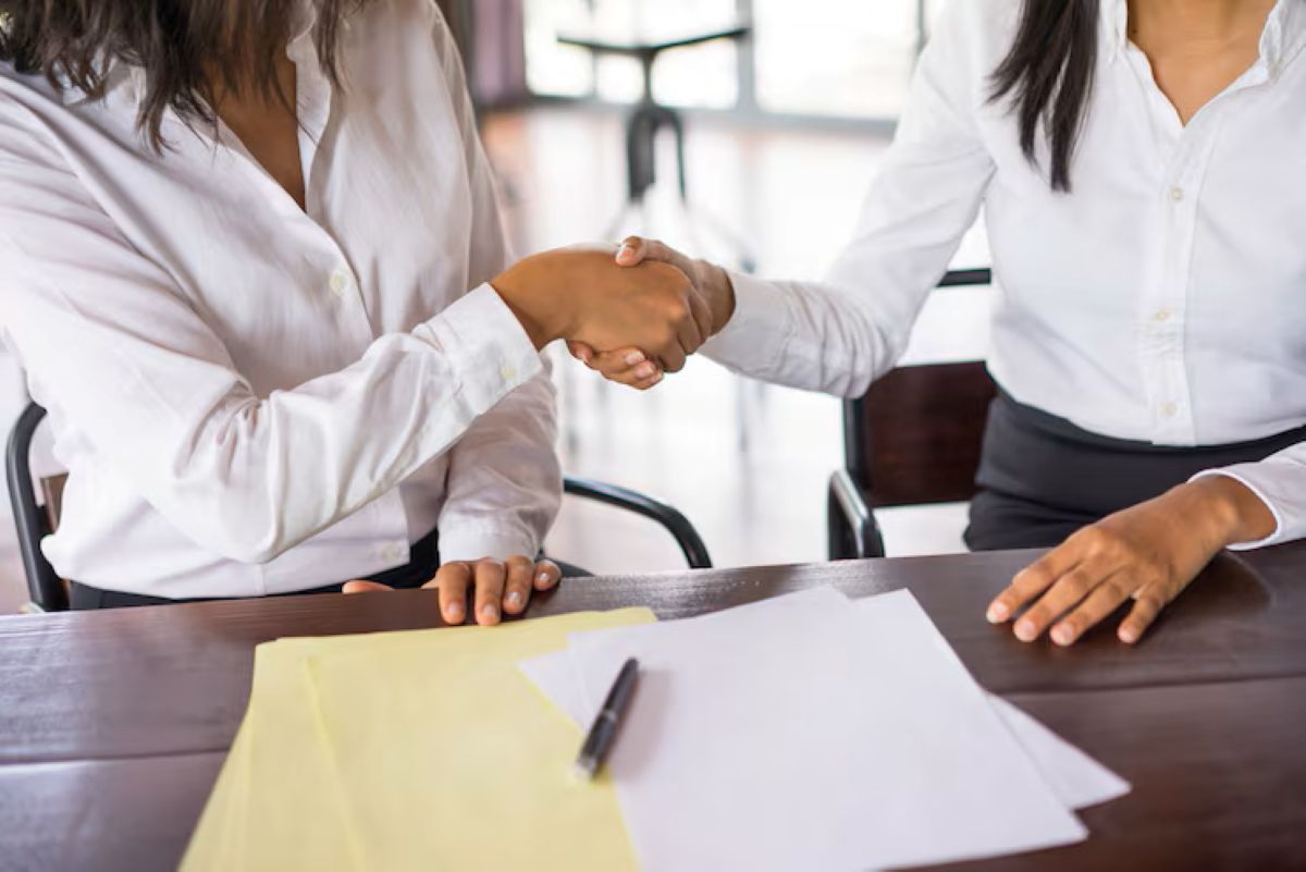 Duas pessoas em camisas sociais brancas apertando as mãos sobre uma mesa de madeira escura com papéis, caneta e documentos, simbolizando a formalização do acerto e o cálculo final da rescisão do contrato de experiência.