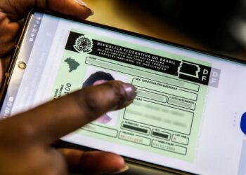Uma mão manuseando um celular que exibe a Carteira Nacional de Habilitação (CNH) Digital. A imagem representa o programa do Governo que oferece cursos gratuitos para tirar a primeira CNH.