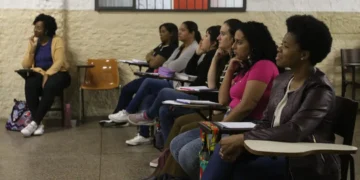 Adultas sentadas em carteiras escolares, prestando atenção em aula em sala comunitária