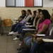 Adultas sentadas em carteiras escolares, prestando atenção em aula em sala comunitária