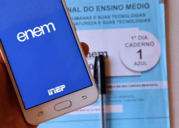 Telefone mostrando tela do ENEM com caderno azul do exame e caneta ao fundo.