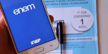 Telefone mostrando tela do ENEM com caderno azul do exame e caneta ao fundo.