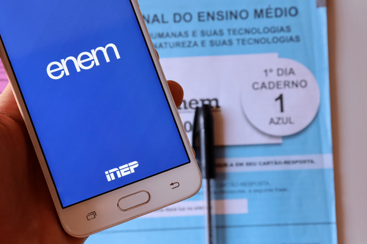 Celular exibindo a tela do ENEM e prova do exame sobre a mesa com caneta.