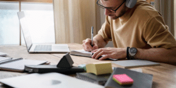 Homem de óculos usando fones de ouvido escrevendo em caderno no escritório com laptop e materiais ao redor
