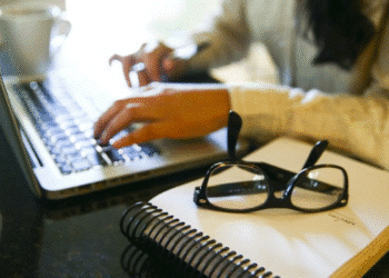Pessoa estudando em casa com notebook, caderno e óculos sobre a mesa.