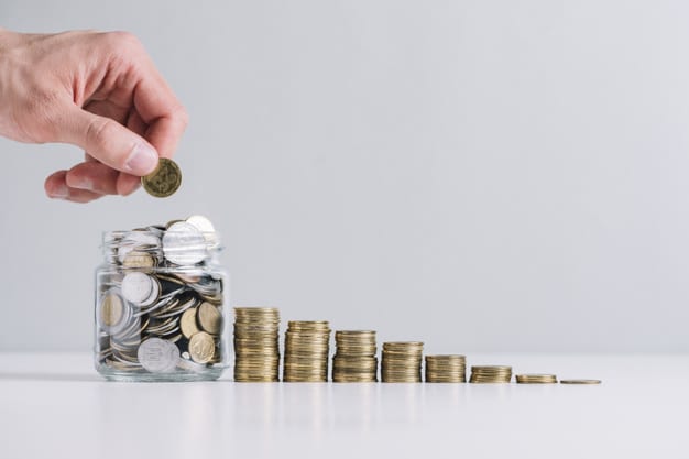 Mão colocando moedas em um pote com outras moedas, simbolizando a prática de economizar dinheiro.