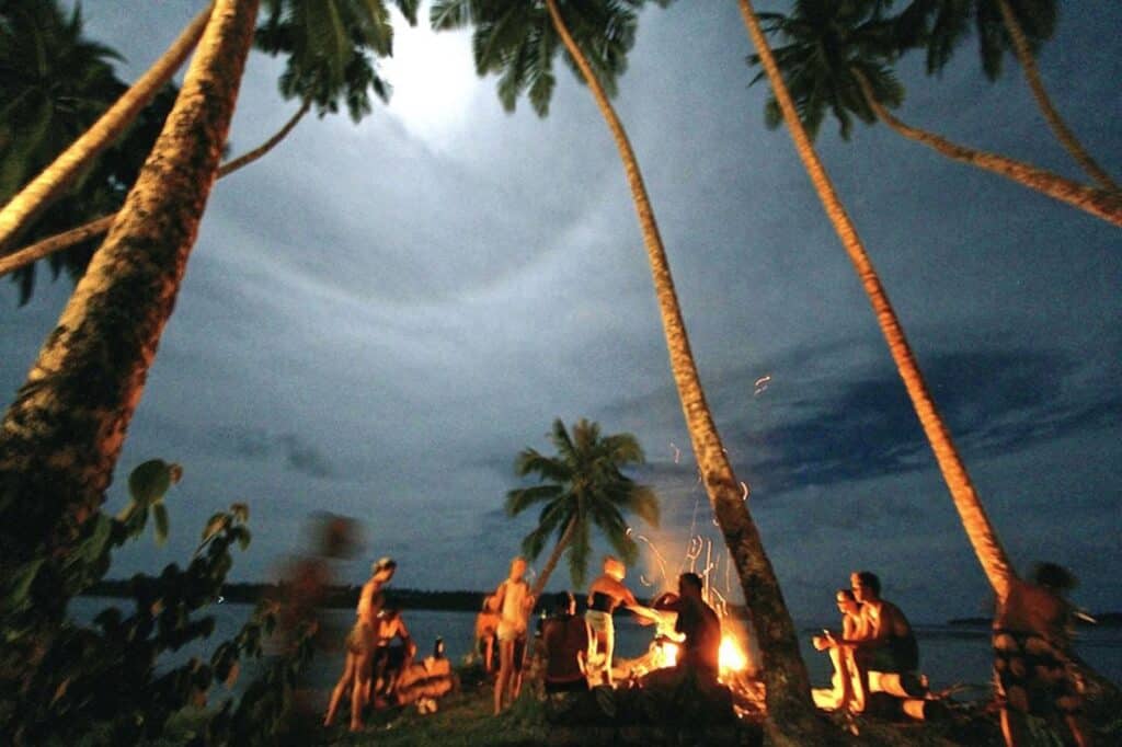 Grupo de pessoas reunidas em um luau à beira-mar sob a lua cheia, com palmeiras e uma fogueira iluminando a noite tropical.