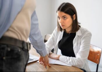 Mulher com semblante sério e preocupado, sentada em uma mesa de escritório, olha para um colega ou superior que está em pé, apontando para um ponto na mesa, em uma situação que sugere uma conversa tensa sobre a demissão durante o período de experiência.