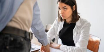 Mulher com semblante sério e preocupado, sentada em uma mesa de escritório, olha para um colega ou superior que está em pé, apontando para um ponto na mesa, em uma situação que sugere uma conversa tensa sobre a demissão durante o período de experiência.