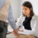 Mulher com semblante sério e preocupado, sentada em uma mesa de escritório, olha para um colega ou superior que está em pé, apontando para um ponto na mesa, em uma situação que sugere uma conversa tensa sobre a demissão durante o período de experiência.