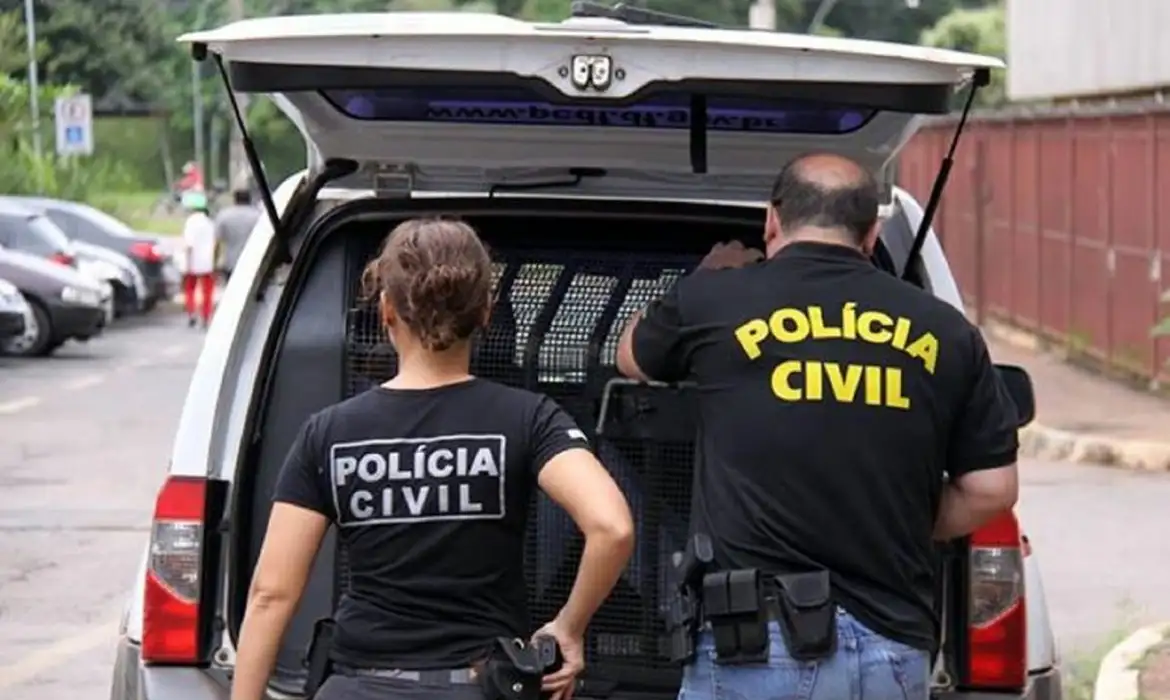 Polícia Civil em ação durante operação de prisão Dois policiais civis, um homem e uma mulher, vestidos com camisetas pretas, em frente a uma viatura aberta