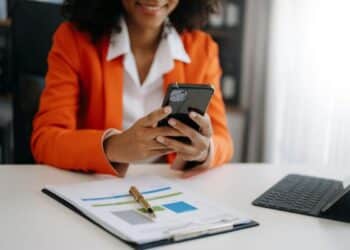 Profissional em blazer laranja usando celular para gerenciamento de tarefas e produtividade com documentos e tablet.