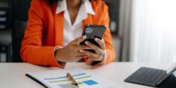 Profissional em blazer laranja usando celular para gerenciamento de tarefas e produtividade com documentos e tablet.