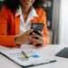 Profissional em blazer laranja usando celular para gerenciamento de tarefas e produtividade com documentos e tablet.
