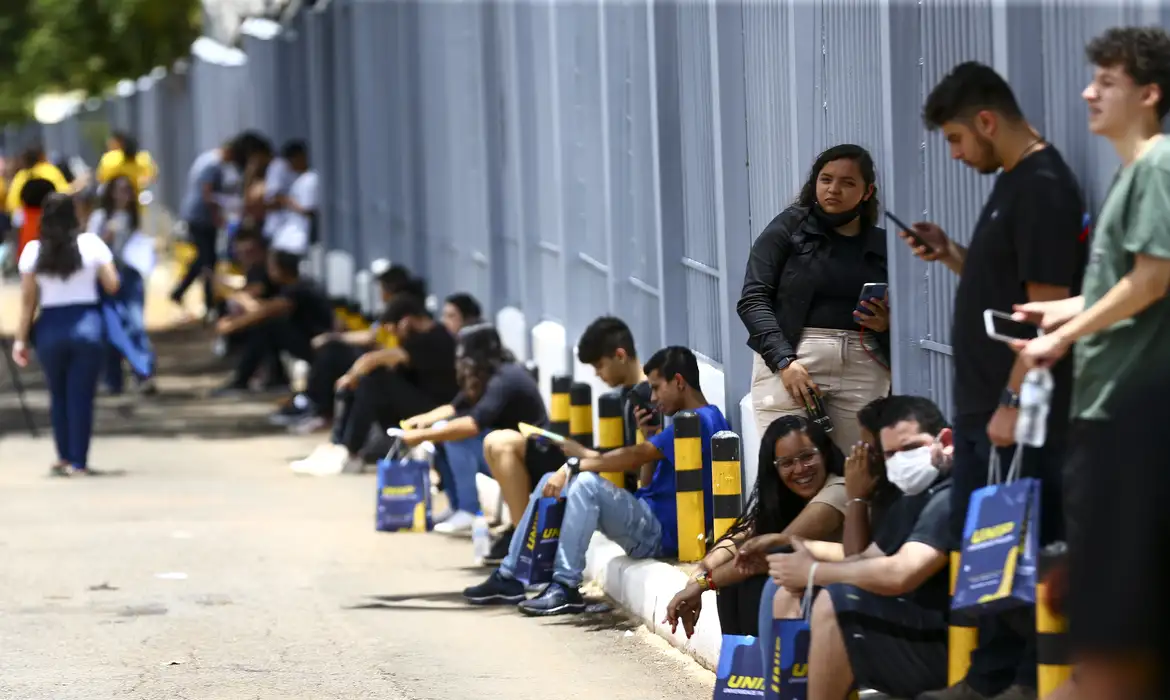 Estudantes aguardam entrada em universidade com mochilas Pessoas jovens sentadas e em pé ao longo de muro, muitas com bolsas da UNIP, aguardando entrada em evento ou aula