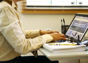 Pessoa usando camisa clara digitando em notebook com óculos e caderno na mesa