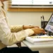 Pessoa usando camisa clara digitando em notebook com óculos e caderno na mesa