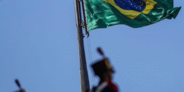Bandeira do Brasil hasteada com céu azul ao fundo e soldado em uniforme cerimonial.