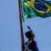 Bandeira do Brasil hasteada com céu azul ao fundo e soldado em uniforme cerimonial.