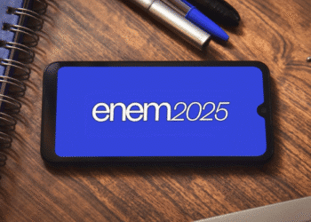 Smartphone exibindo o texto "Enem2025" em tela azul sobre mesa de madeira com caderno, canetas e plugue.