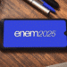 Smartphone exibindo o texto "Enem2025" em tela azul sobre mesa de madeira com caderno, canetas e plugue.