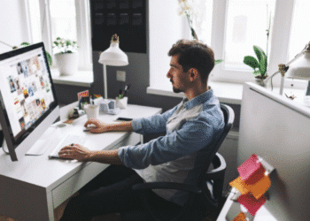 Homem trabalhando como designer gráfico em um computador, com a tela exibindo diversas imagens e projetos visuais, em um ambiente de escritório claro e moderno.