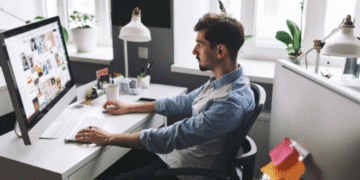Homem trabalhando como designer gráfico em um computador, com a tela exibindo diversas imagens e projetos visuais, em um ambiente de escritório claro e moderno.