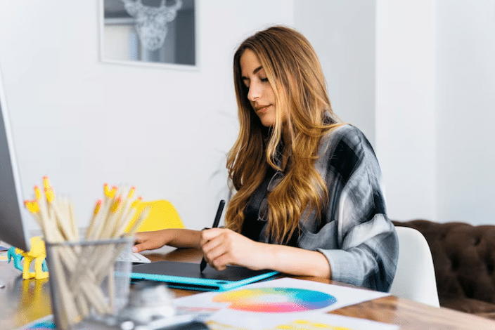 Mulher trabalhando como designer gráfico, utilizando uma mesa digitalizadora e caneta em sua mesa de trabalho, com um círculo cromático e lápis de cor por perto.