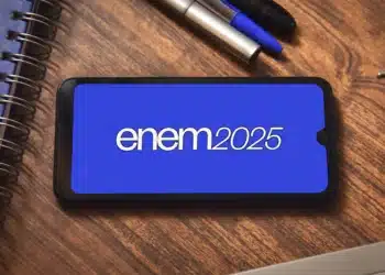 Um smartphone exibe a inscrição 'enem 2025' em sua tela azul, simbolizando a preparação para o exame. O aparelho está sobre uma mesa de madeira, ao lado de um caderno e canetas.