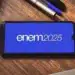 Um smartphone exibe a inscrição 'enem 2025' em sua tela azul, simbolizando a preparação para o exame. O aparelho está sobre uma mesa de madeira, ao lado de um caderno e canetas.