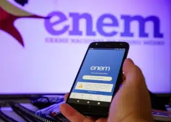 Pessoa segurando celular com aplicativo do ENEM aberto na tela e logo do ENEM ao fundo