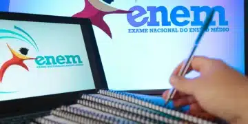 Mão segurando uma caneta sobre cadernos espirais, com um laptop ao fundo exibindo o logo do ENEM (Exame Nacional do Ensino Médio), simbolizando os estudos para a prova.