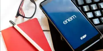 Smartphone com tela mostrando logo do ENEM, ao lado de teclado, caderno vermelho, lápis e óculos.