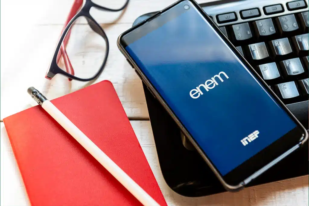 Acesso Digital e Comunicação do ENEM 2025: Nota Oficial do Ministro Smartphone com o logo do ENEM e Inep sobre um teclado de computador. Próximo, há um caderno vermelho, um lápis e um par de óculos de leitura.