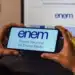 Mãos segurando um celular com o logotipo "ENEM - Exame Nacional do Ensino Médio" em primeiro plano, sobre um notebook que exibe a mesma logo.