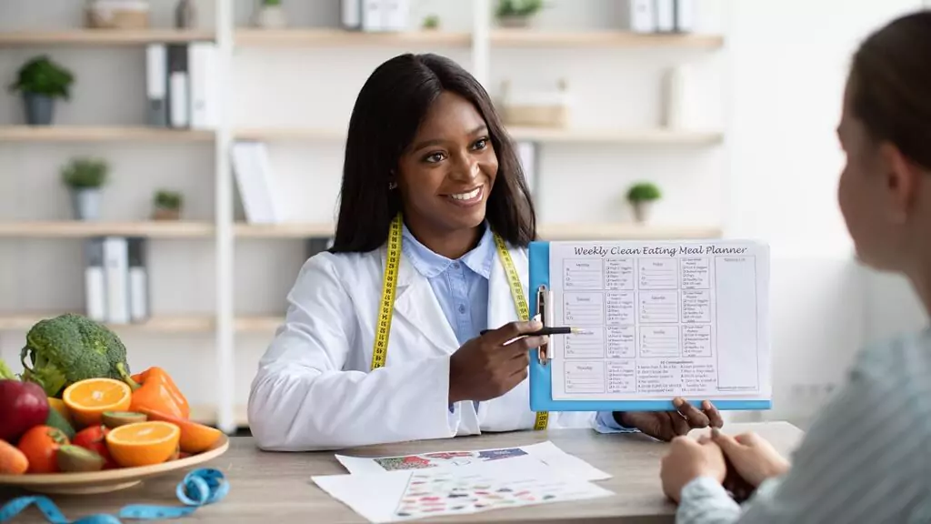 Orientação Nutricional: Promovendo Hábitos Saudáveis para o Bem-Estar Nutricionista sorridente explicando um plano de alimentação saudável a um paciente, com mesa cheia de frutas e vegetais, representando a ascensão da saúde preventiva.
