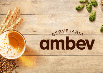 Logo da Cervejaria Ambev ao lado de um copo de cerveja, grãos de cevada e lúpulo sobre superfície de madeira