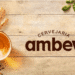 Logo da Cervejaria Ambev ao lado de um copo de cerveja, grãos de cevada e lúpulo sobre superfície de madeira